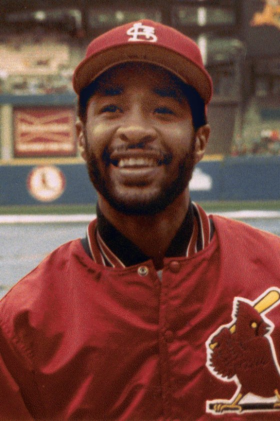 Ozzie Smith smiling in a St. Louis Cardinals cap and red Cardinals warm-up jacket on the field at Busch Memorial Stadium, 1983